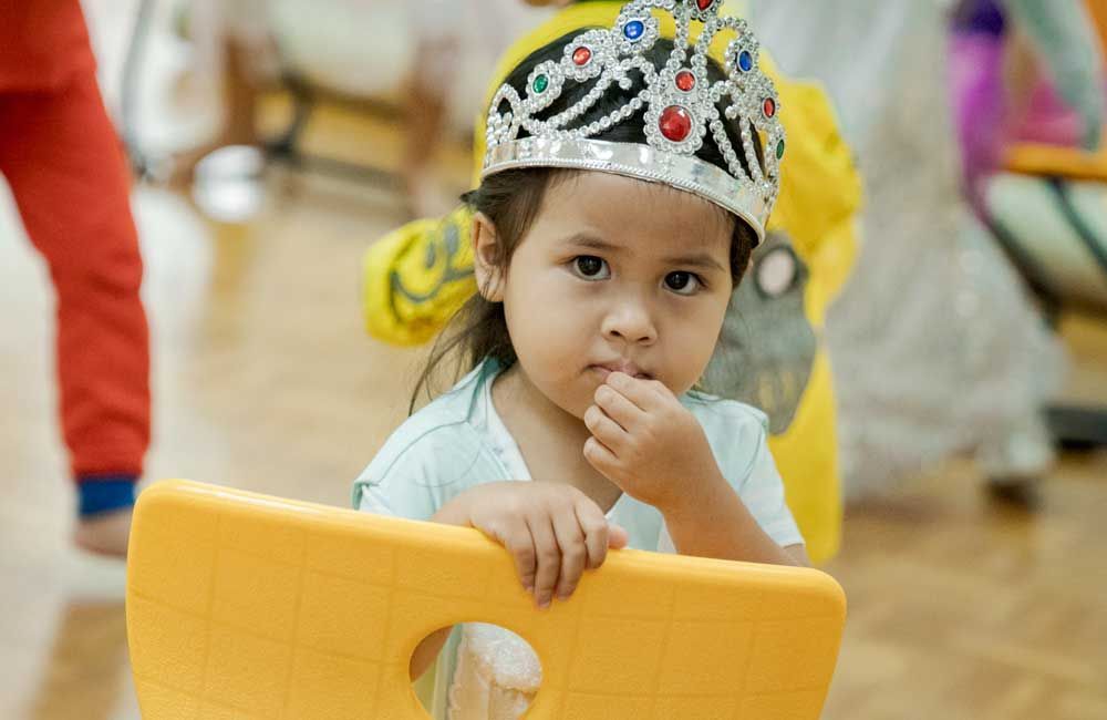 A little girl wearing a crown is sitting on a yellow chair.
