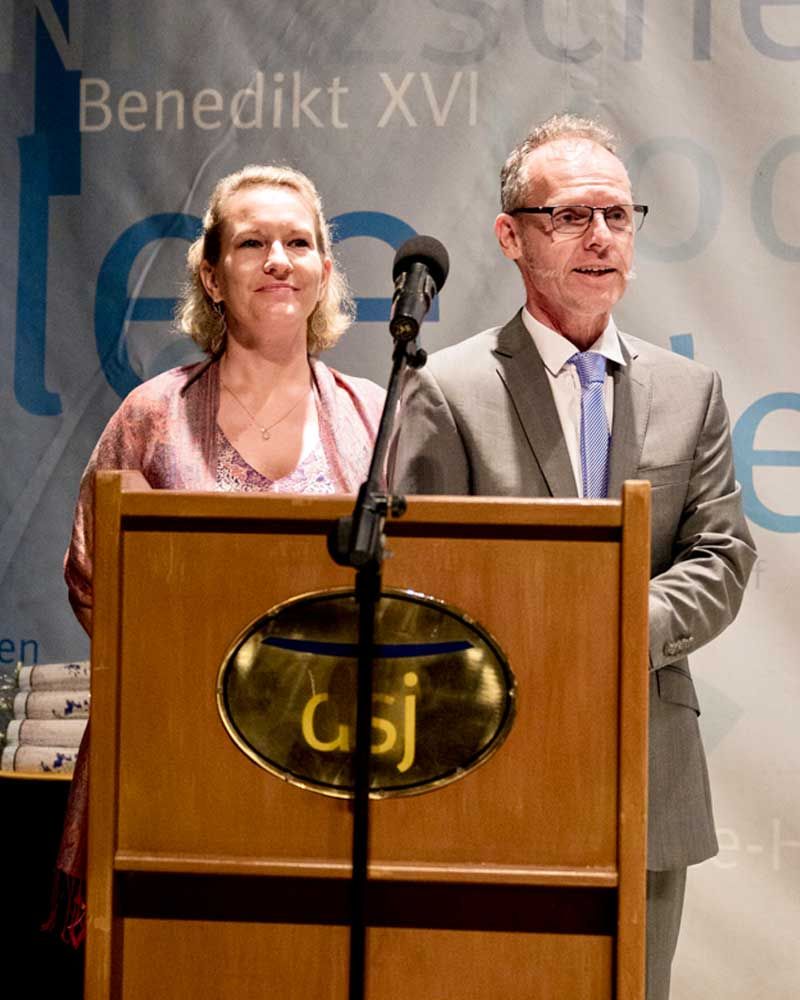 A man and a woman stand at a podium with benedikt xvi in the background