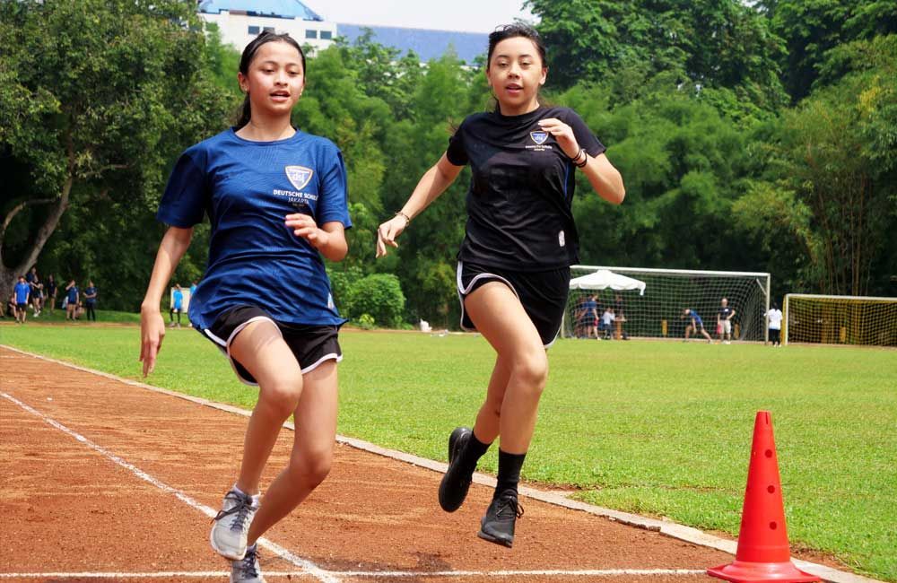 Two girls are running on a track next to a red cone.