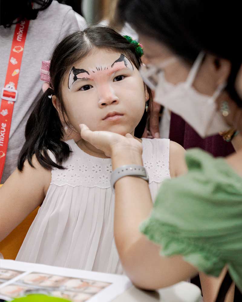 A little girl is getting her face painted by a woman wearing a mask