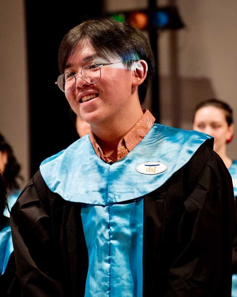 A young man wearing glasses and a blue gown is smiling.