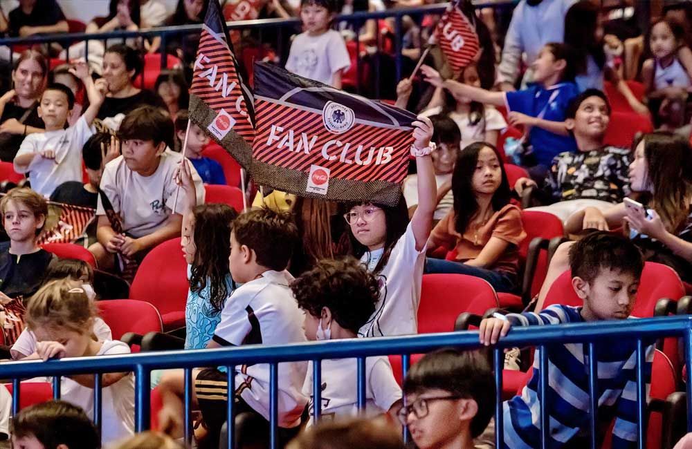 A group of children are sitting in a stadium holding a fan club flag.