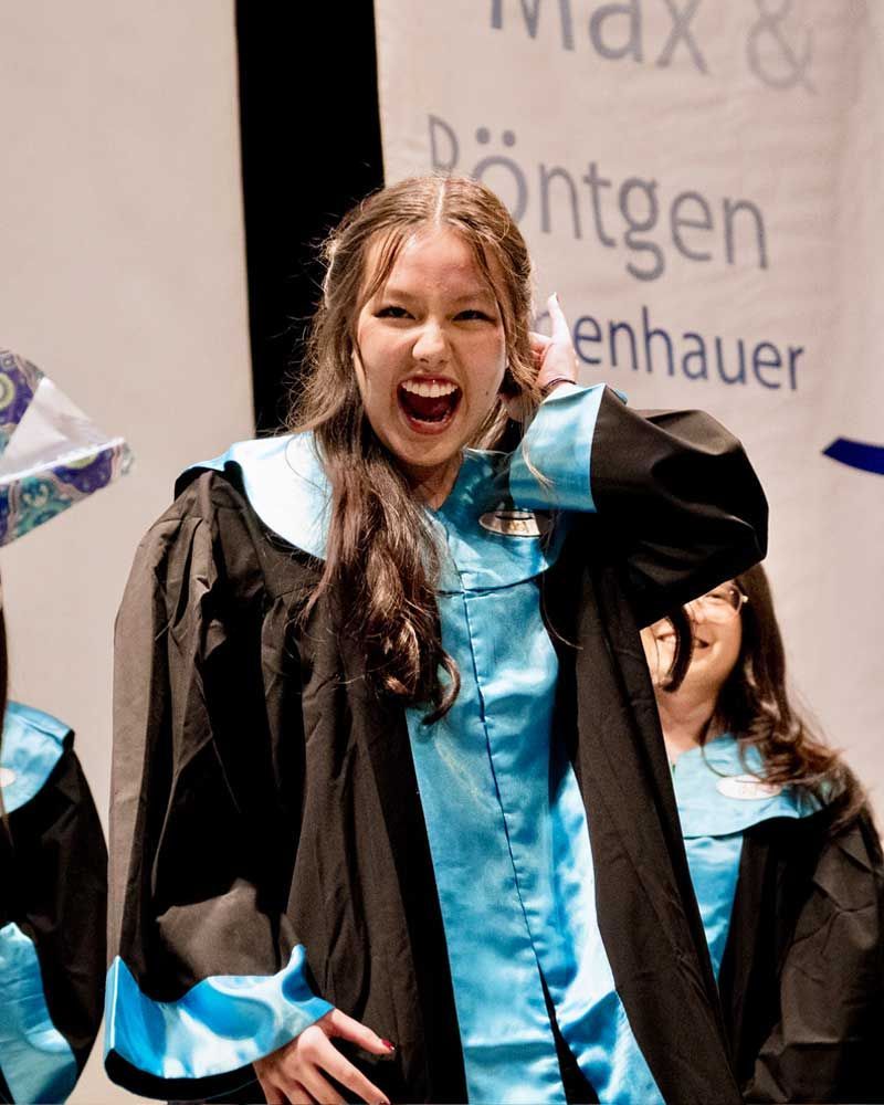 A woman in a graduation gown stands in front of a sign that says max
