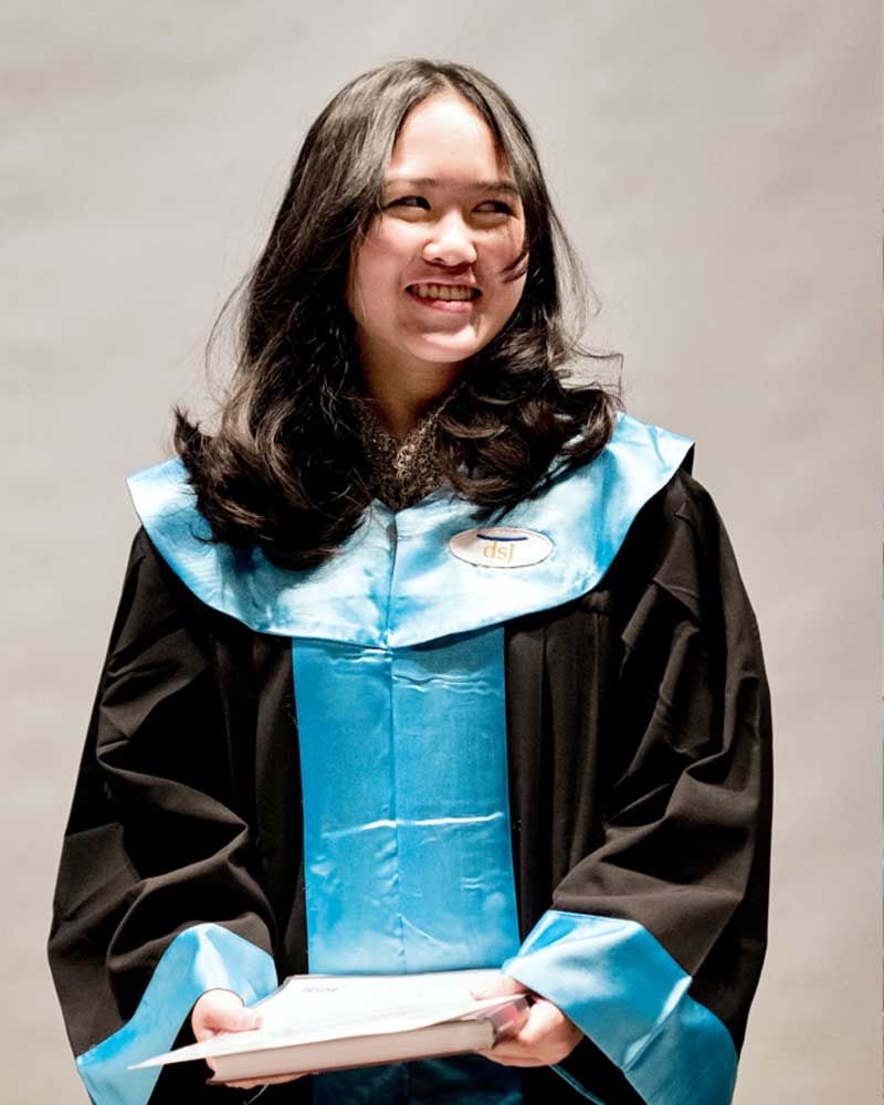 A woman in a graduation gown is smiling and holding a piece of paper