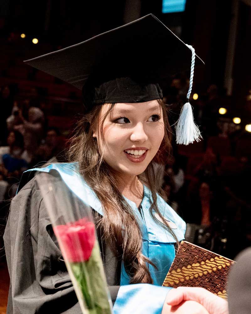 A woman in a graduation cap and gown is holding a red rose.