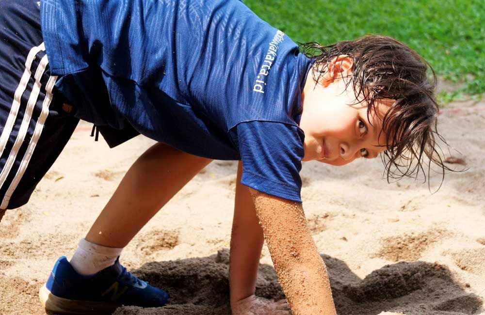 A young boy in a blue shirt is doing push ups in the sand.