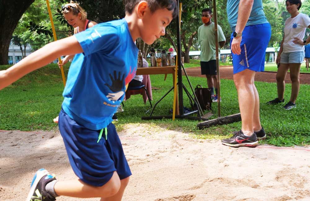 A young boy in a blue shirt is jumping in the sand.