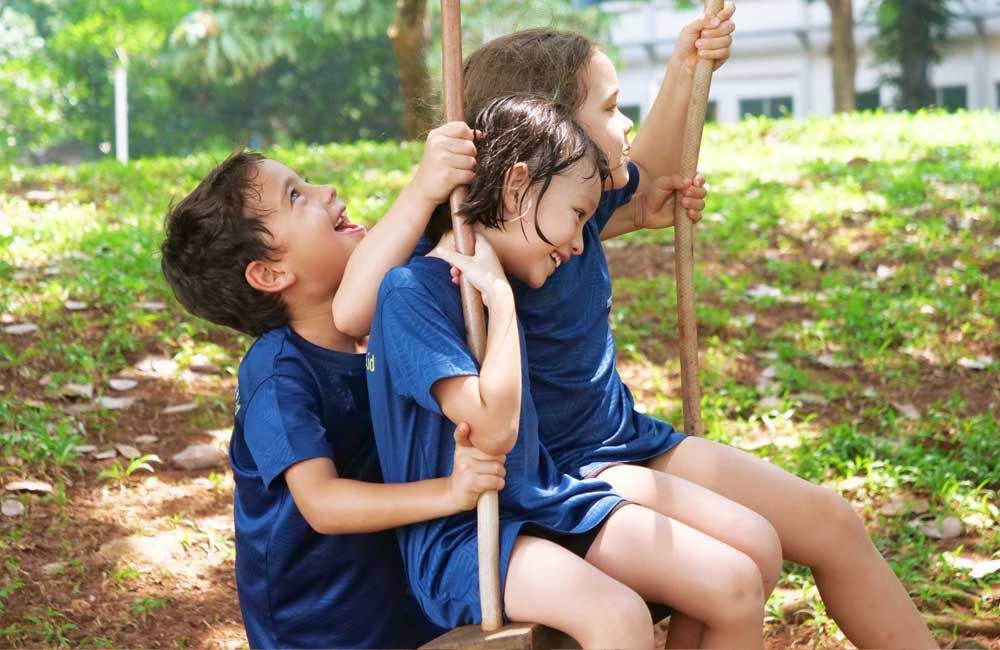 Three children are sitting on a wooden swing in a park.