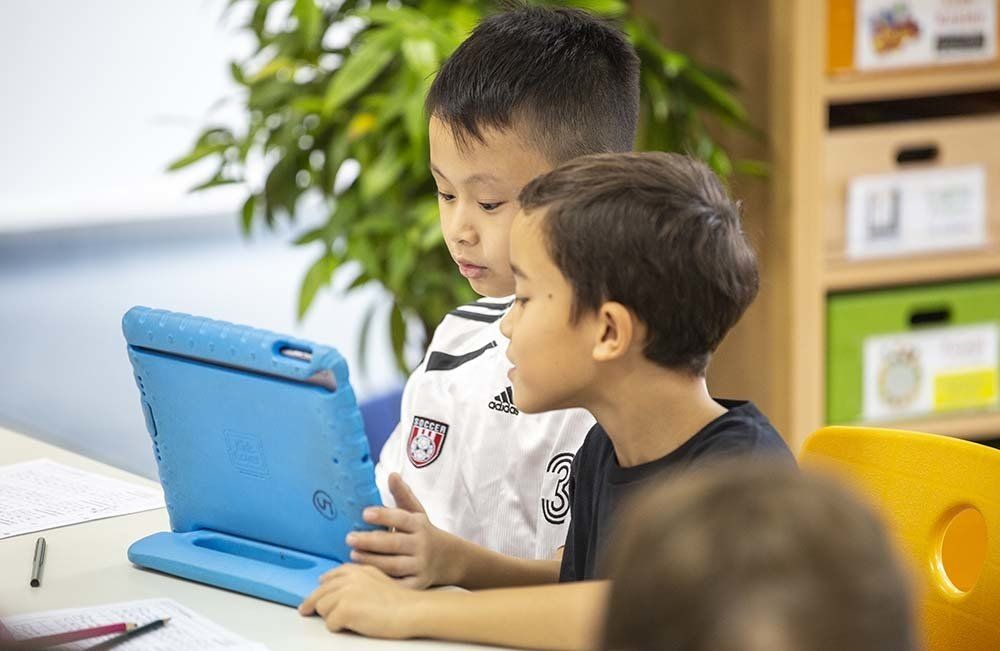 Two young boys are sitting at a table looking at a tablet computer.