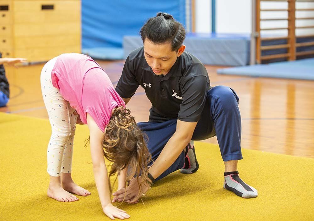 A man is helping a little girl do a handstand in a gym.