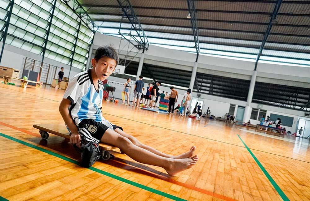 A young boy is sitting on a skateboard in a gym.