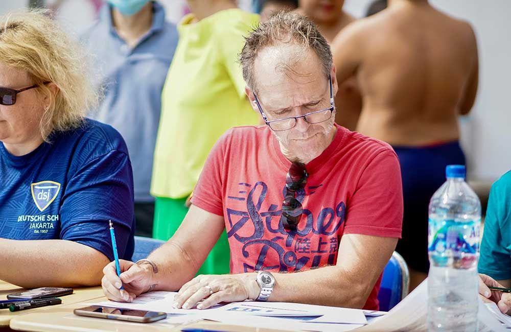 A man in a red shirt is sitting at a table writing on a piece of paper.