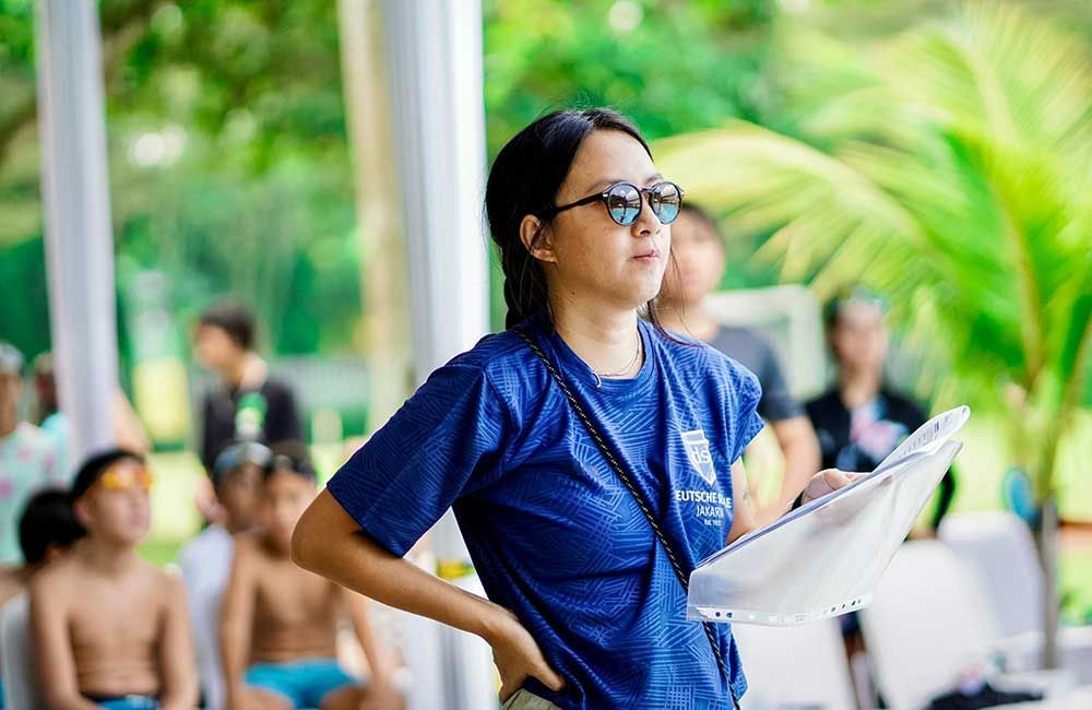 A woman in a blue shirt and sunglasses is standing in front of a group of children.