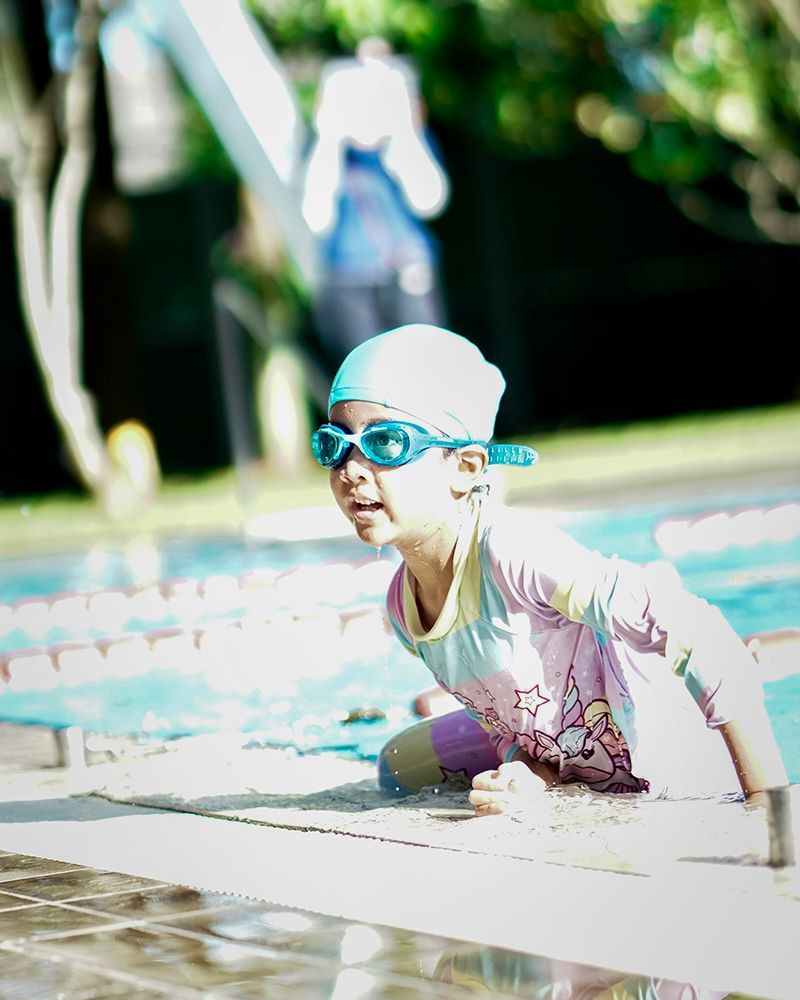 A little girl wearing goggles and a swim cap is crawling into a swimming pool.