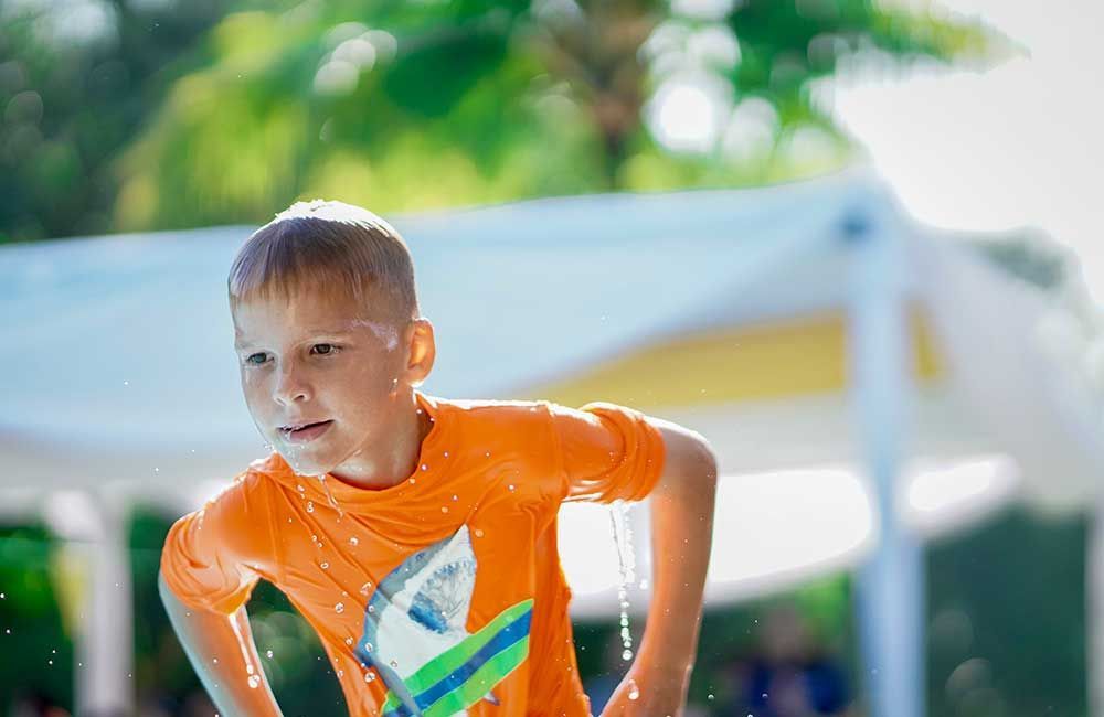 Boy in an orange shirt, soaked with water, standing near a pool with a joyful expression.