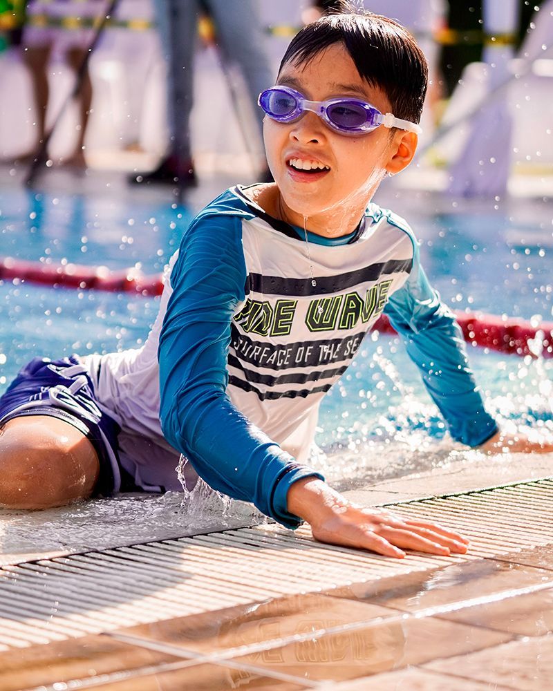 A young boy wearing goggles is laying on the edge of a swimming pool.