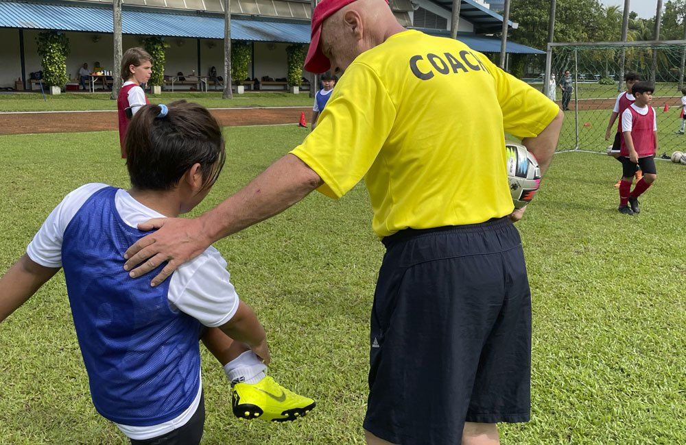 A man in a yellow shirt with the word coach on the back is talking to a child on a soccer field.