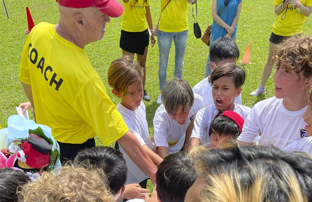 A man in a yellow coach shirt is talking to a group of children.