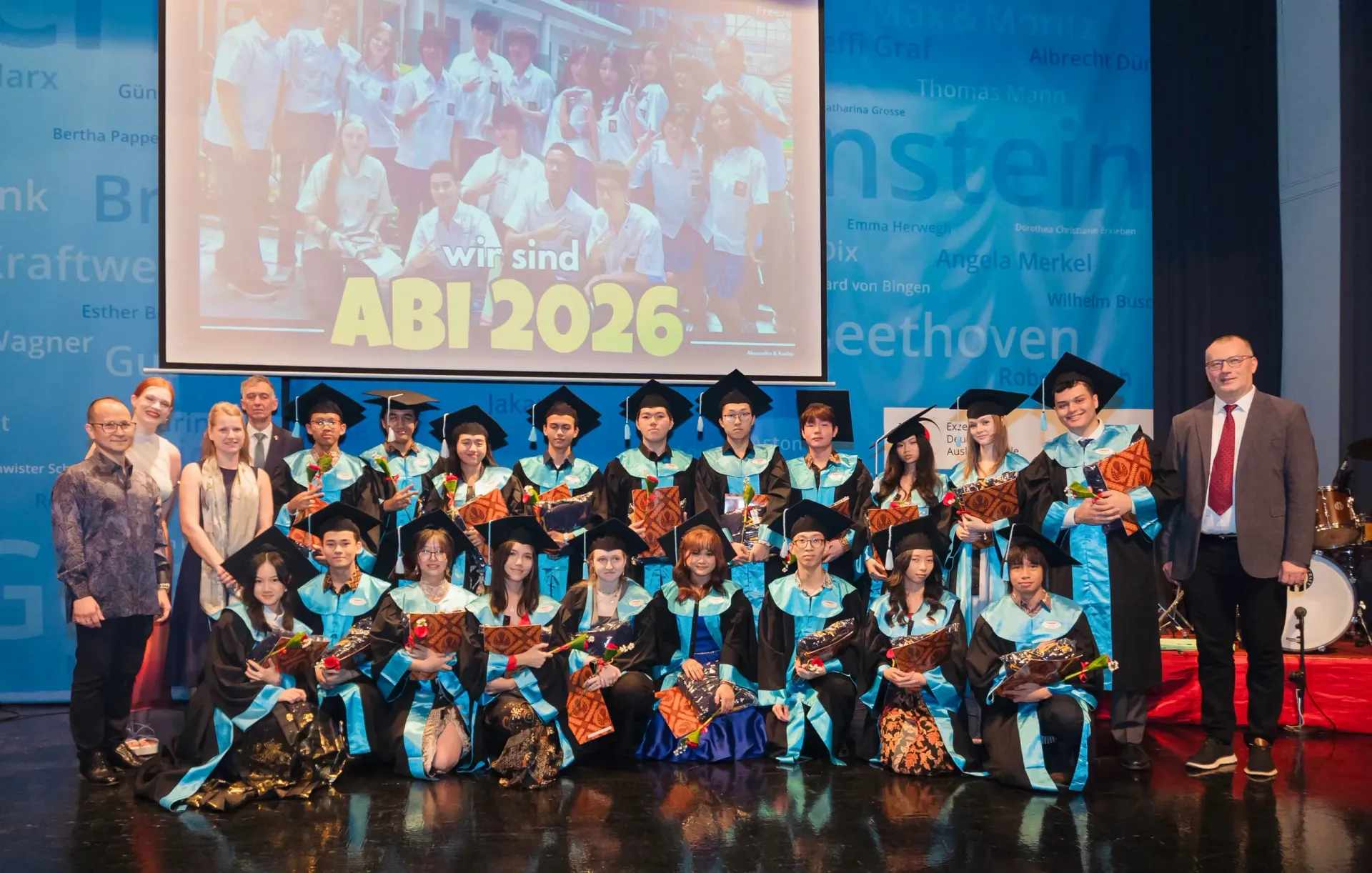 Group of graduates in blue graduation gowns on stage with a large “ABI 2026” screen behind them