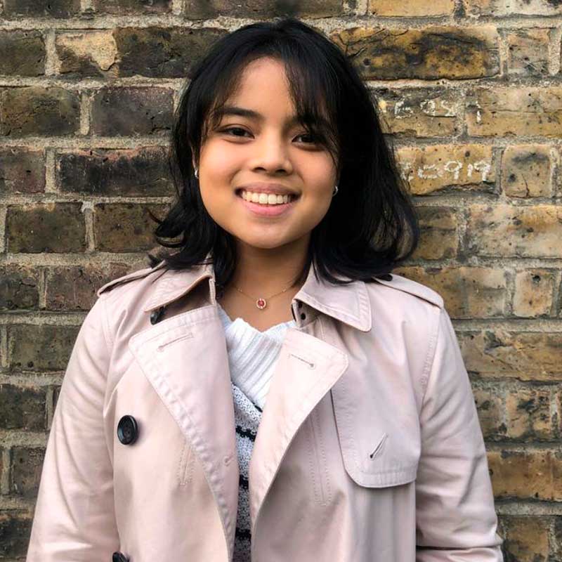 A woman in a trench coat is smiling in front of a brick wall.