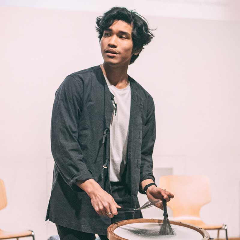 A man in a black jacket is playing a drum with a brush.