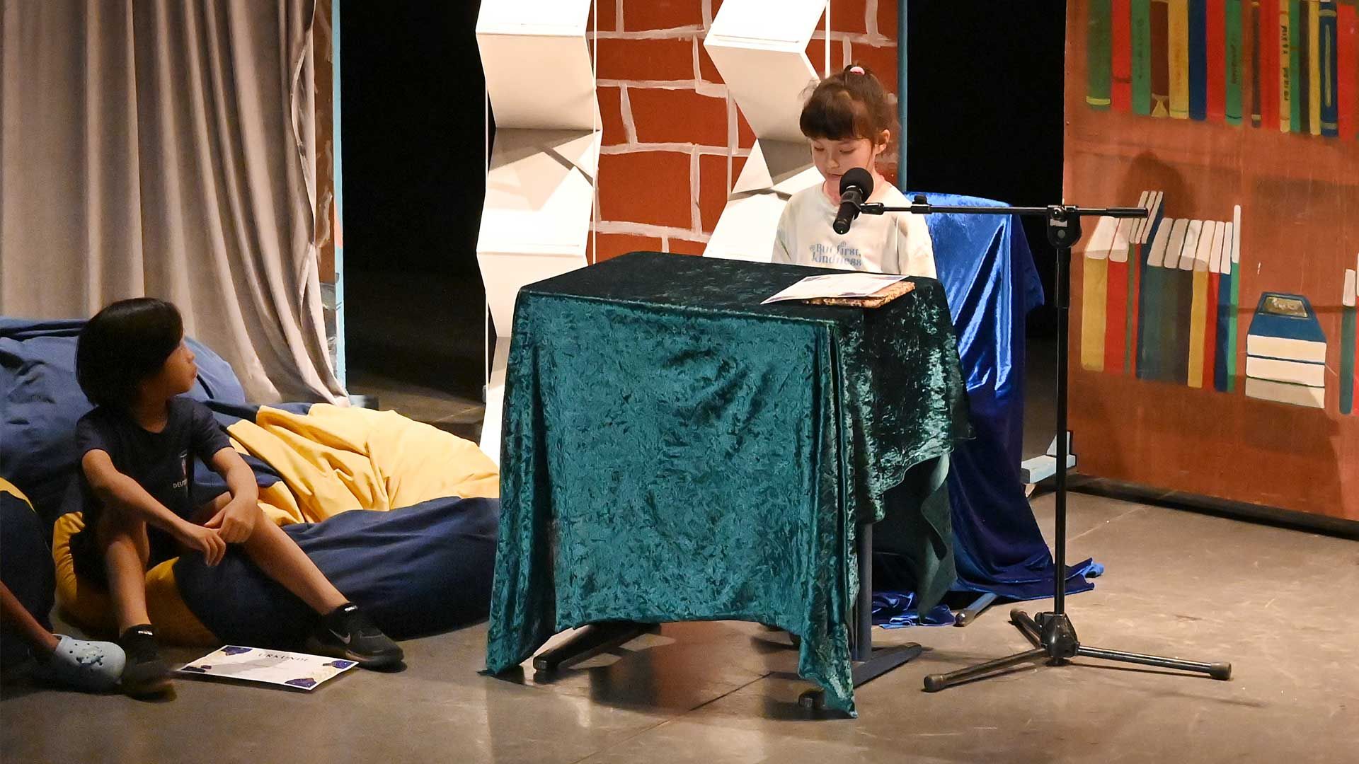 A boy is sitting at a table talking into a microphone while another boy looks on.