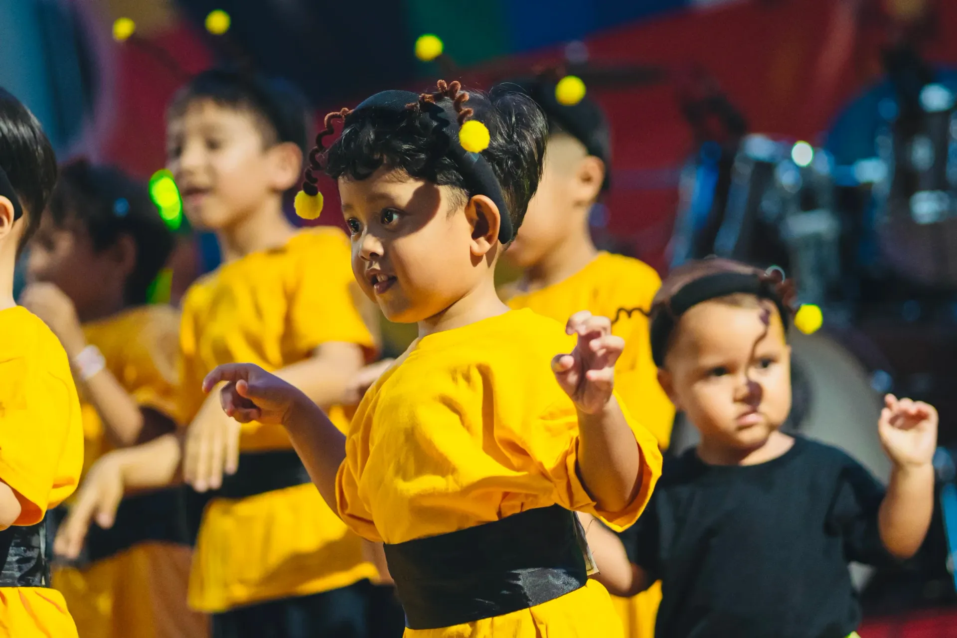 Children in yellow costumes dancing on a stage with colorful confetti and blurred lights in the background