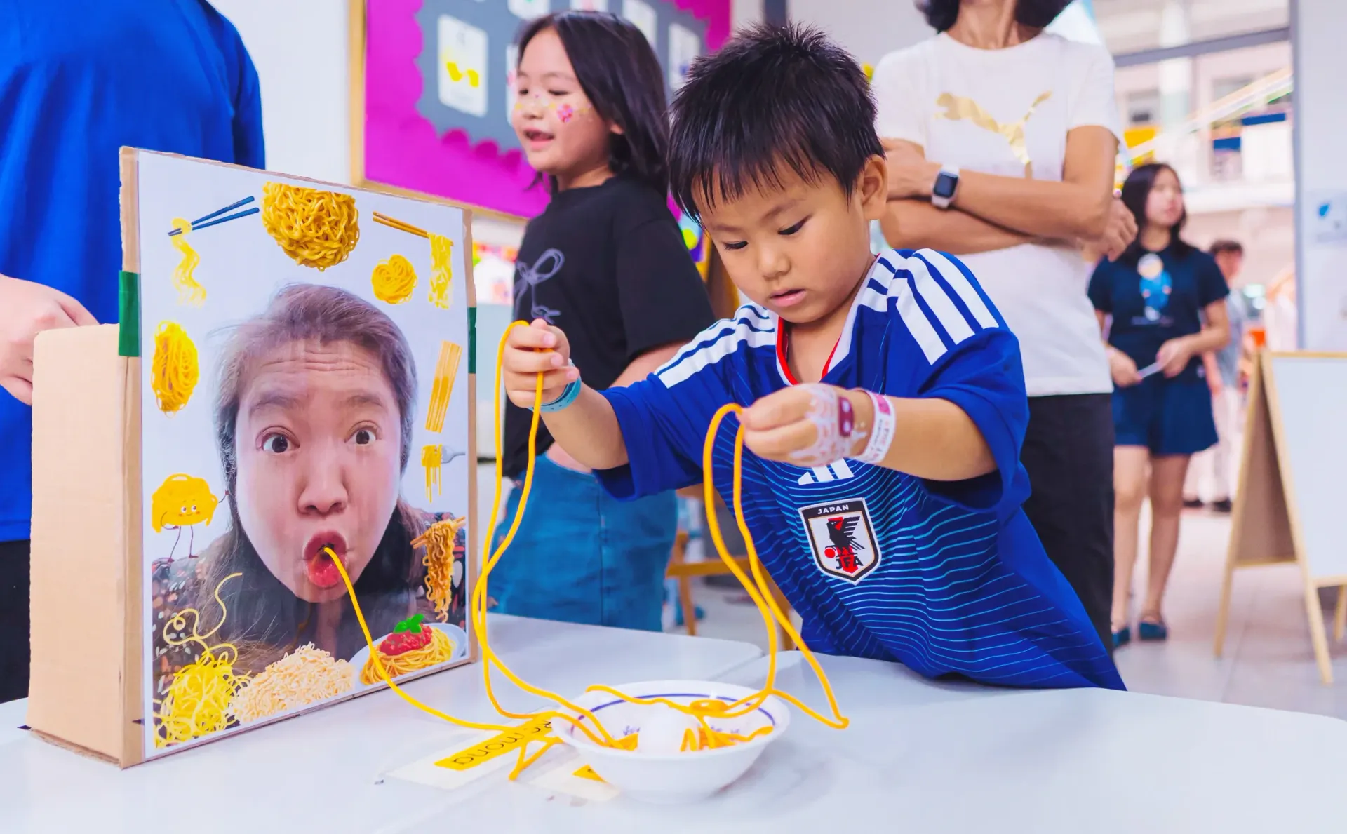 Child threading yellow strings through a board with a face cutout at a colorful indoor event