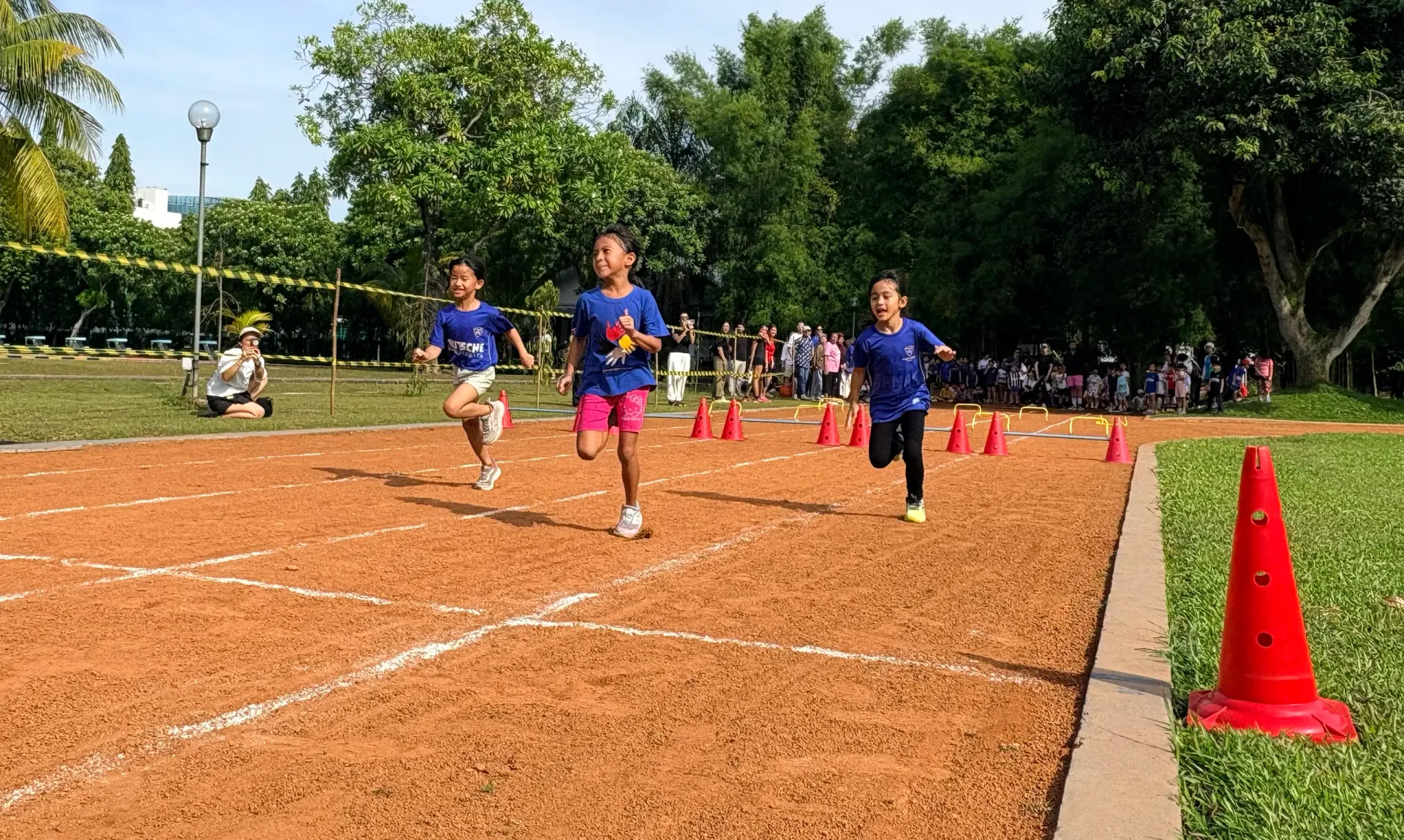 Children running a race on a dirt track, with cones and spectators in a park setting.