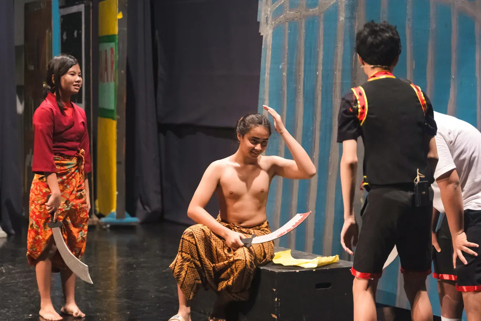 Theater scene with a shirtless performer seated center, others standing nearby on stage under bright lights.