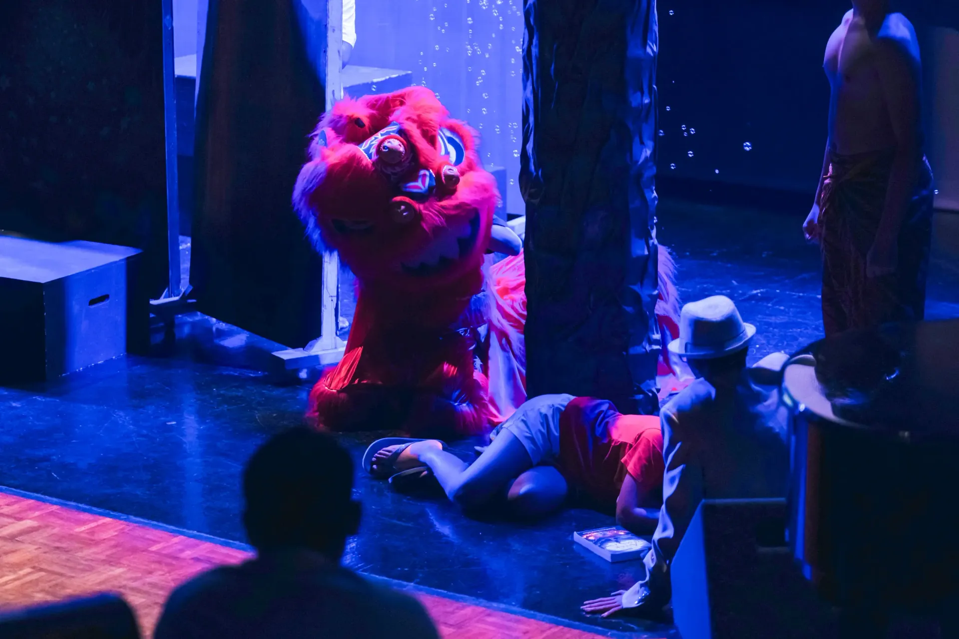 Red puppet or performer on a stage, with a person lying on the floor under blue and red lighting.