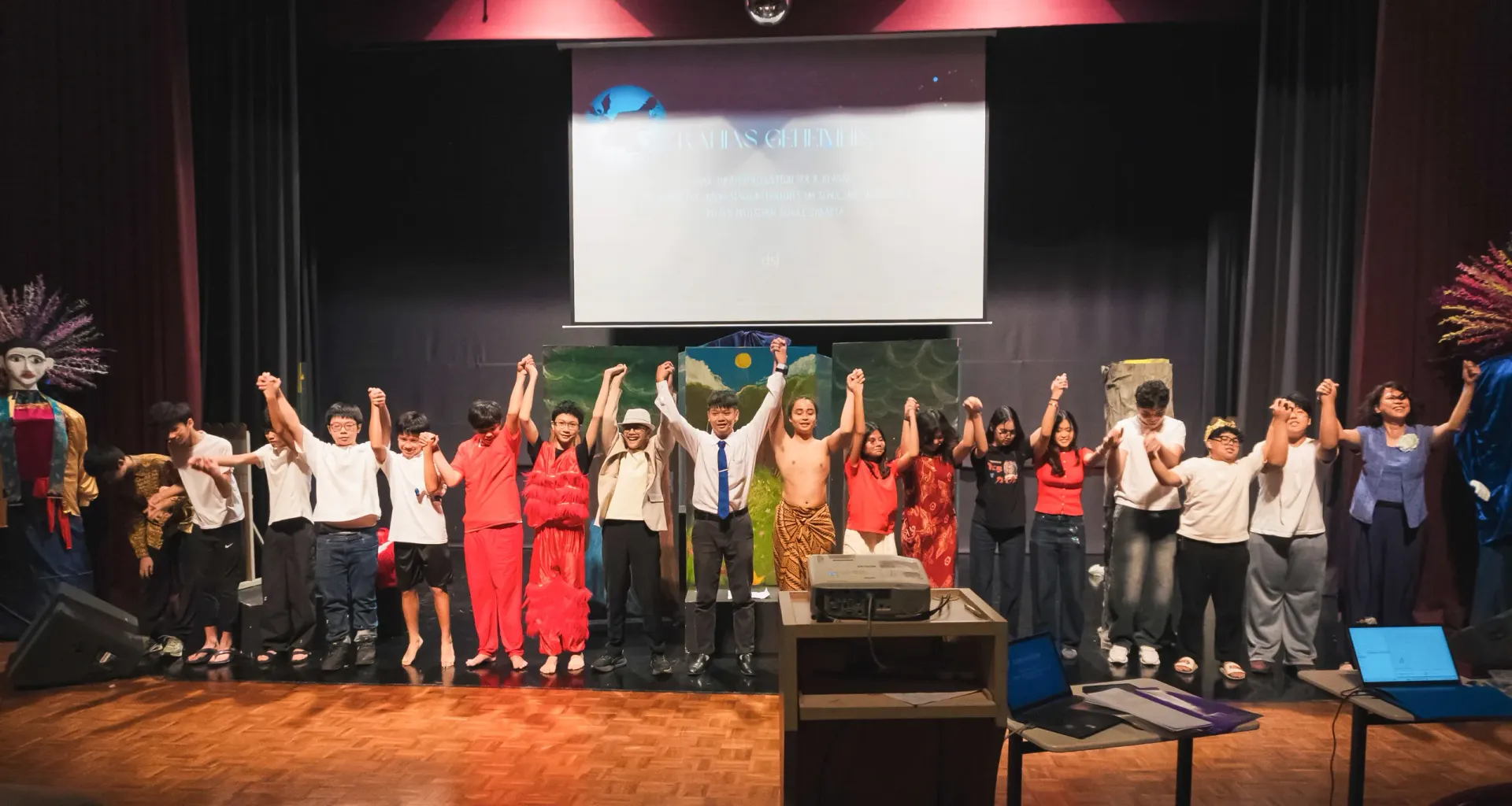 Group of people on a stage with raised hands during a presentation, under a projection screen