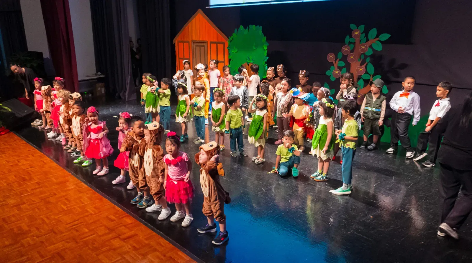 Children in colorful costumes performing on a theater stage with a village set backdrop