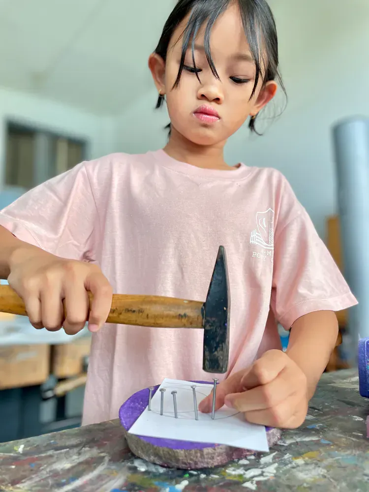 Young girl in a pink shirt using a hammer to work on a craft project, with a focused expression.