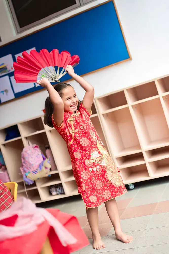 A little girl in a red dress is holding a red fan in a classroom.