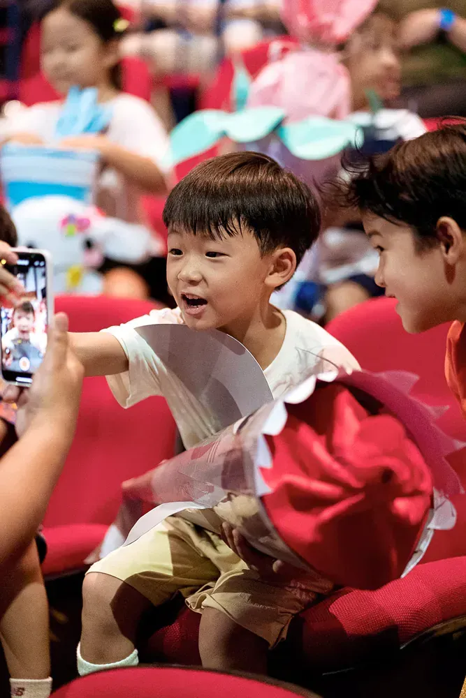 A little boy is sitting in a chair while a person takes a picture of him with a cell phone.
