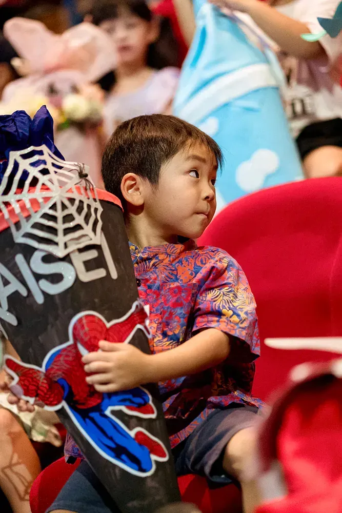 A young boy is sitting in a red chair holding a spiderman sign.