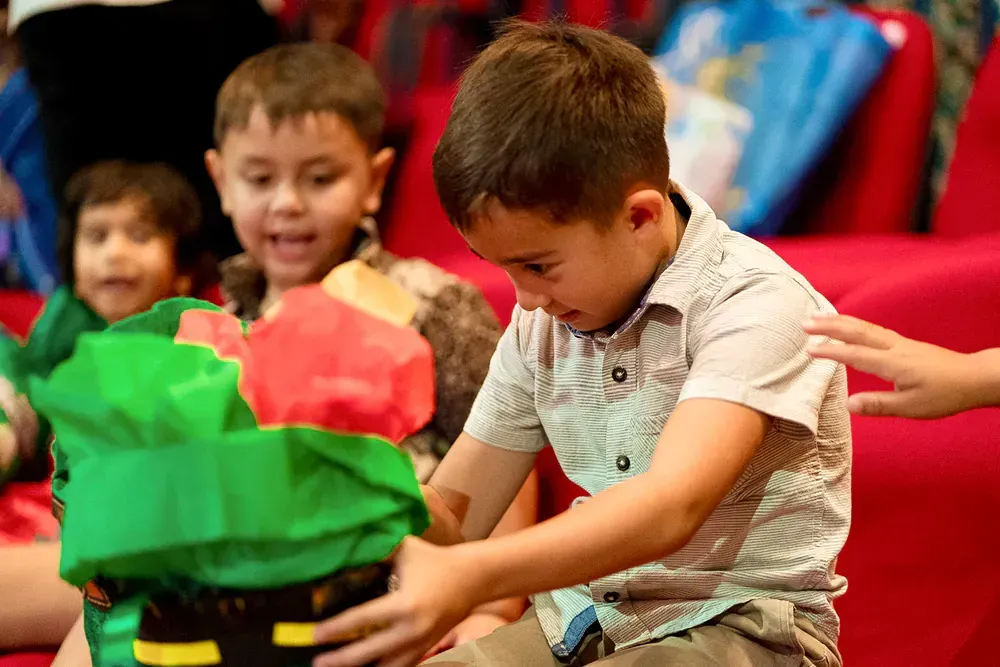 A young boy is opening a gift box while sitting on a red couch.