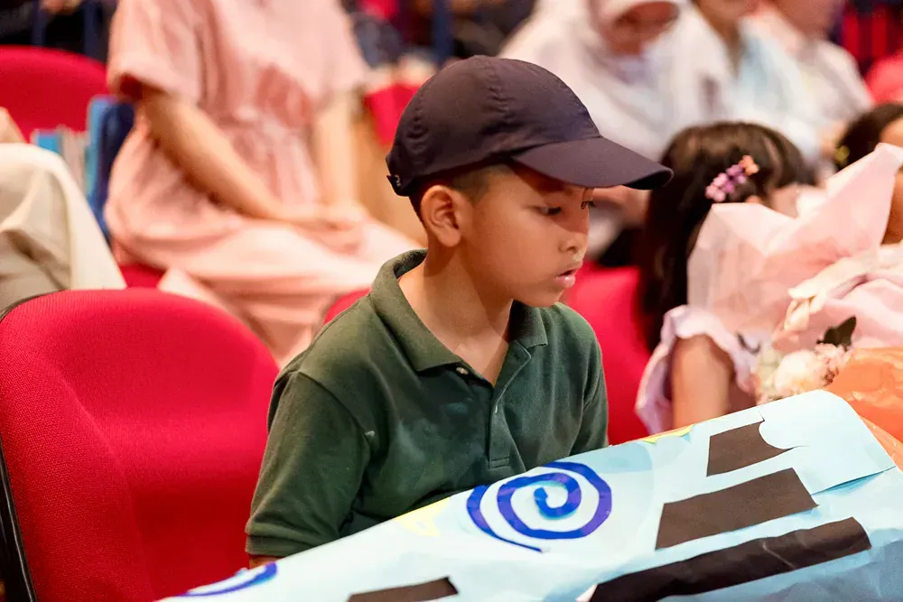 A young boy wearing a hat is sitting in a theatre holding a bag.