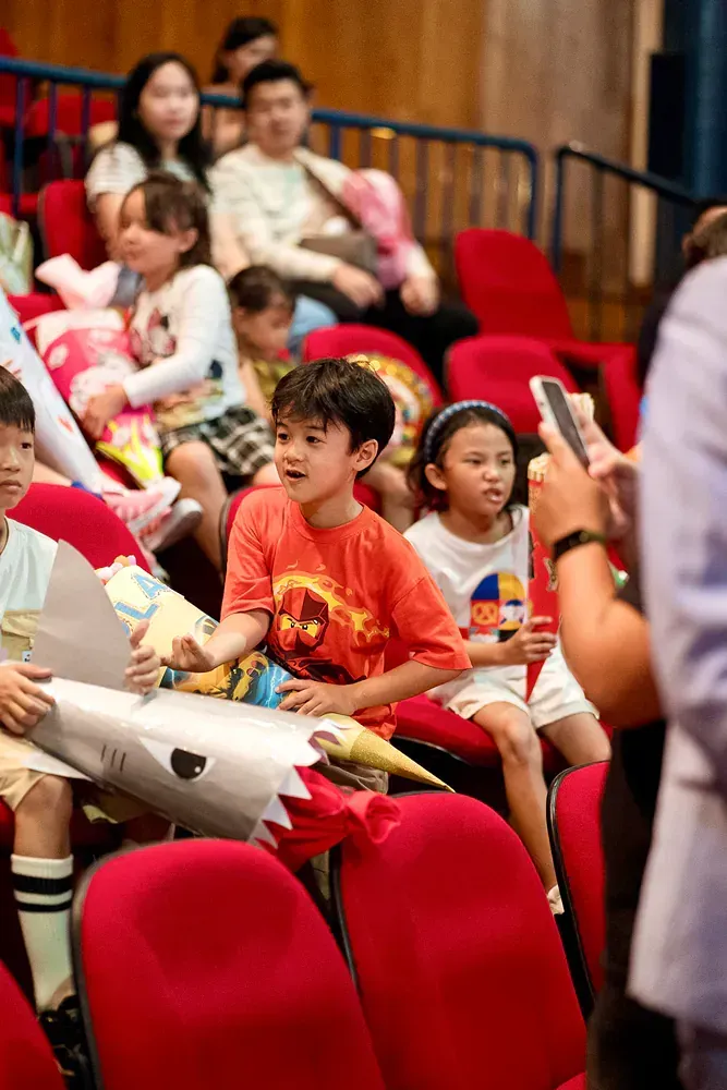 A group of children are sitting in red chairs in a theater.
