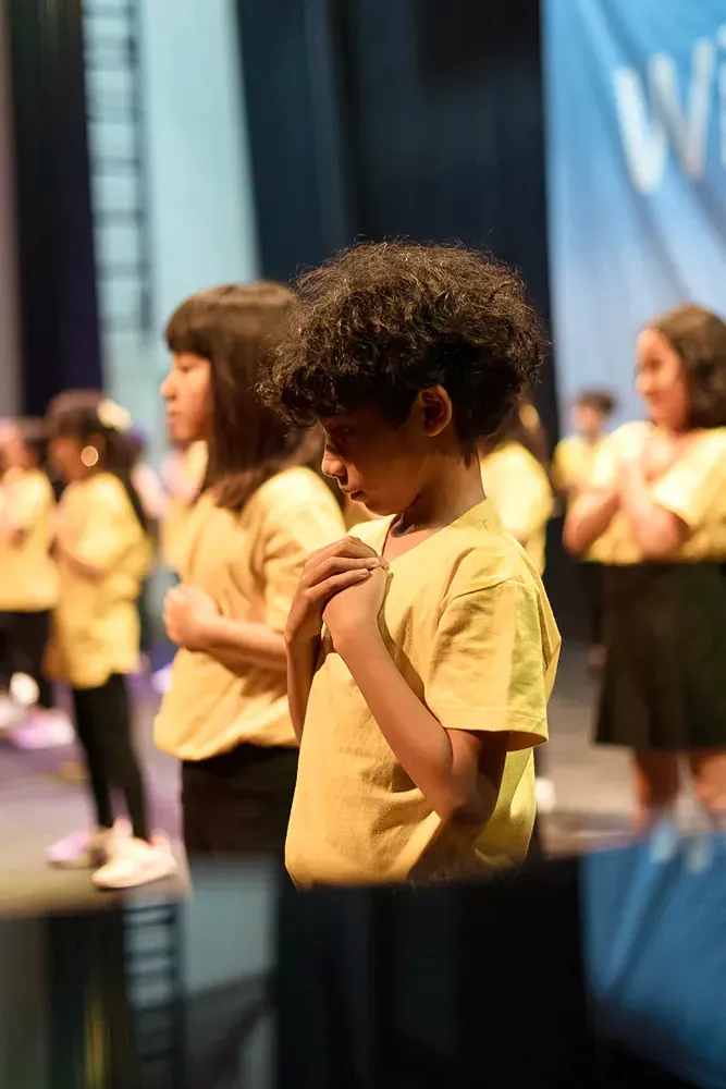 A group of children are standing on a stage with their hands folded in prayer.