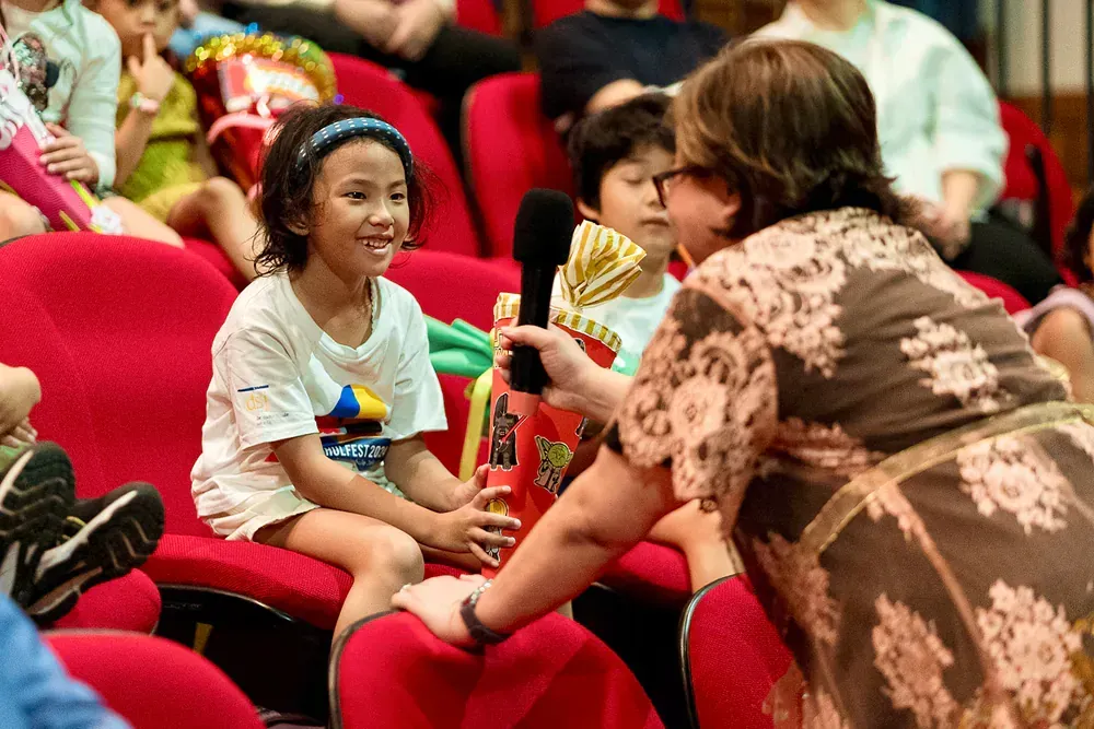 A woman is holding a microphone in front of a group of children sitting in red chairs.