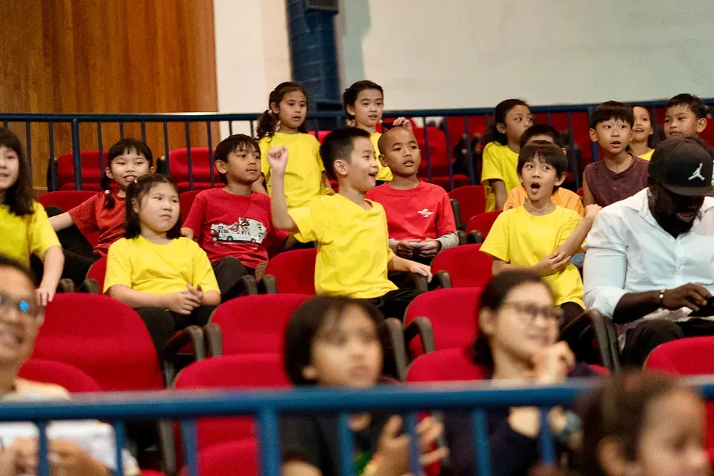 A group of children are sitting in red chairs in an auditorium.
