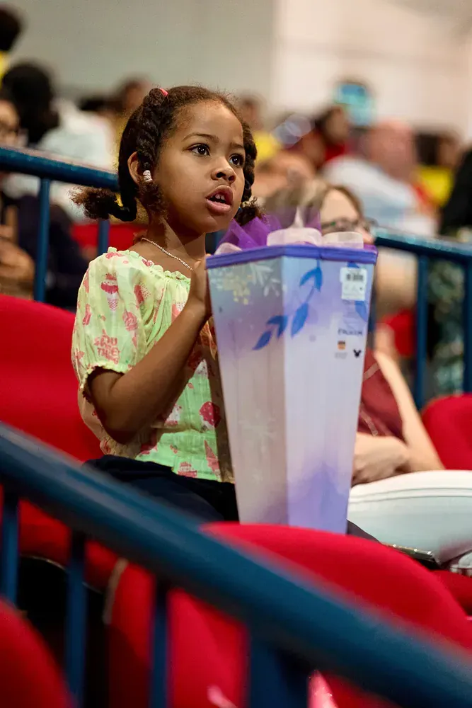A little girl is sitting in a stadium holding a purple container.