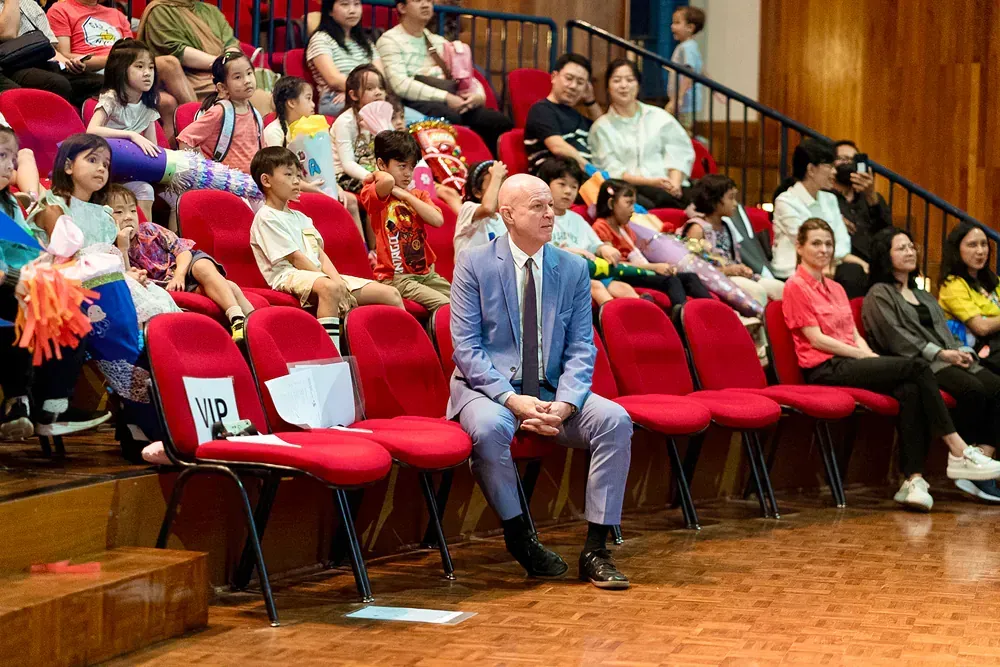 A man in a suit is sitting in a row of red chairs in front of a crowd of people.