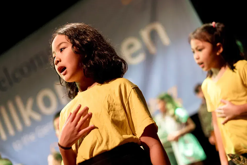 A girl in a yellow shirt is singing in front of a sign that says willkommen