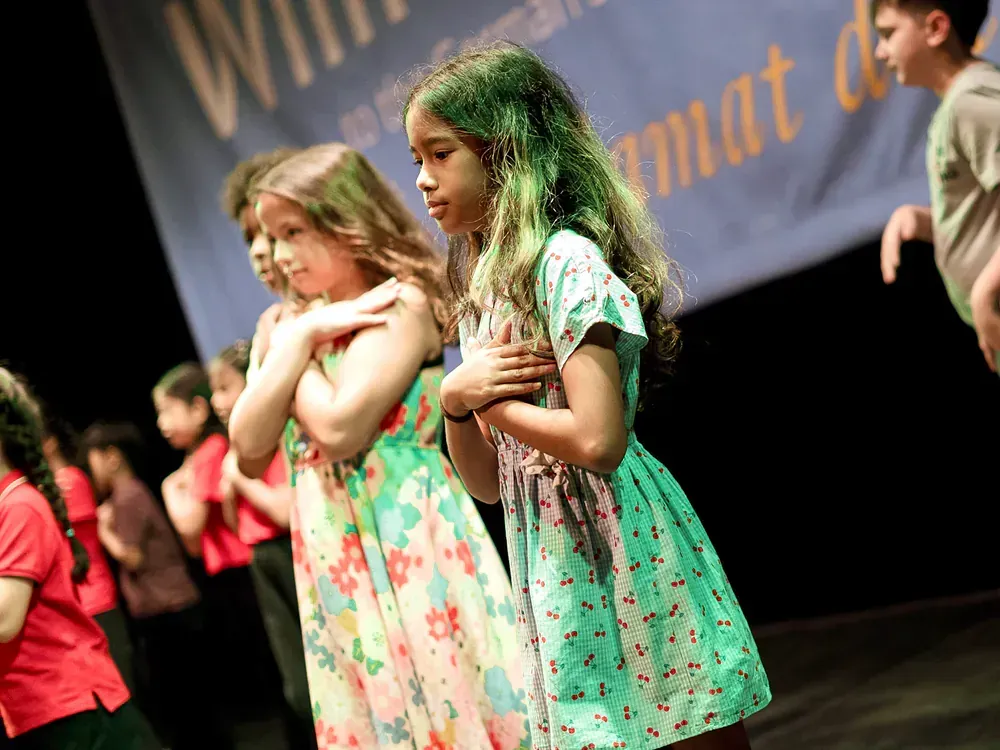 A group of young girls are standing on a stage with their hands folded in front of a banner that says