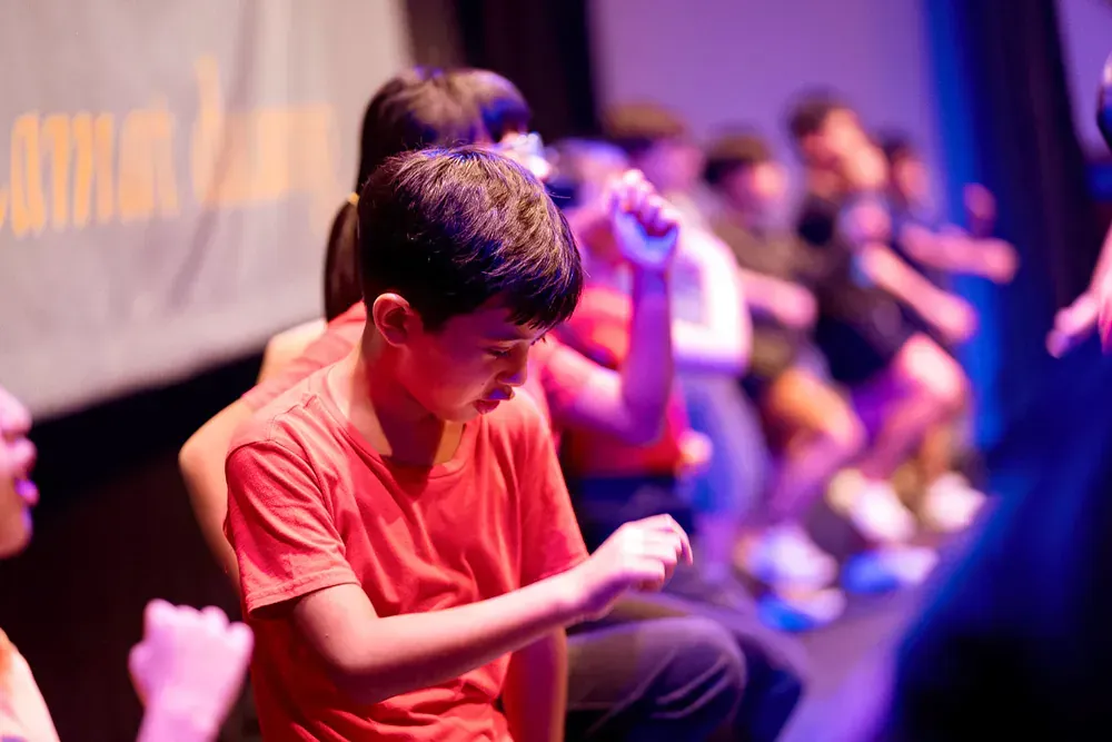 A boy in a red shirt is sitting on a stage in front of a crowd.