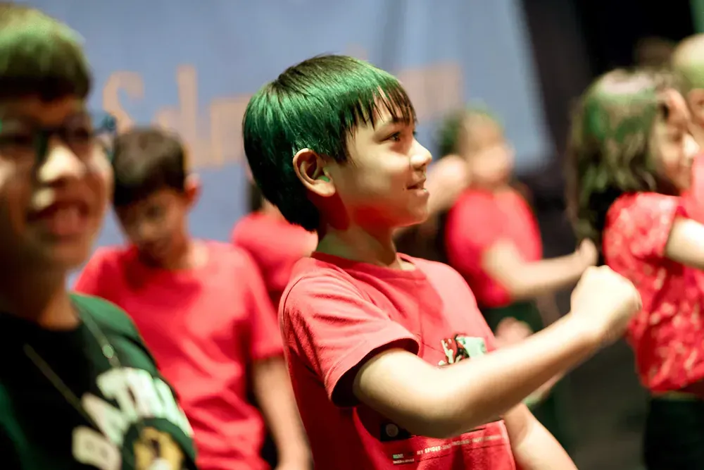 A boy with green hair is standing in front of a group of children in red shirts.