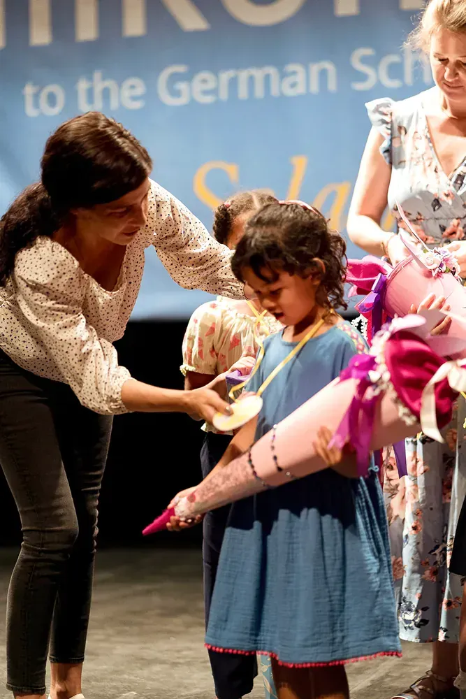 A woman is giving a medal to a little girl in front of a sign that says german school